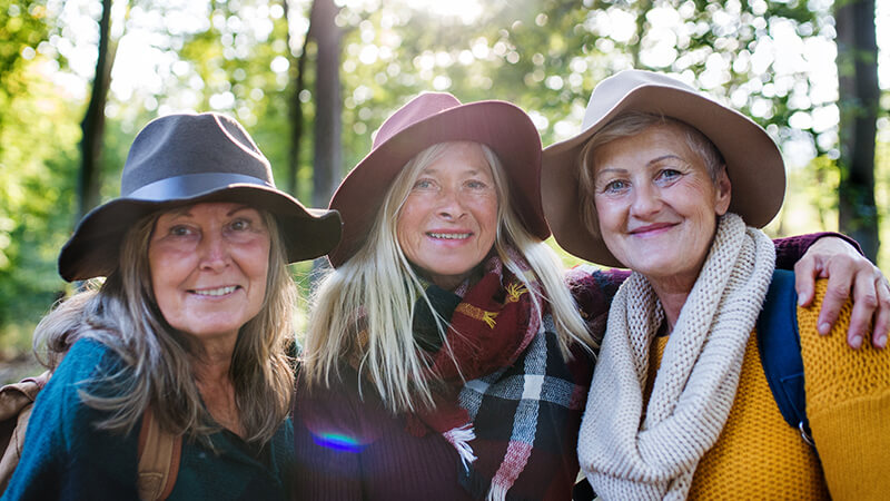 Drei ältere Freundinnen mit Sonnenhüten im Wald lächeln in die Kamera.
