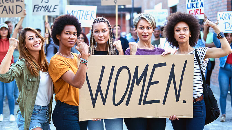 Fünf Frauen zeigen Stärke bei einem Protest für Frauenrechte und halten ein Schild mit dem Wort “Women”.