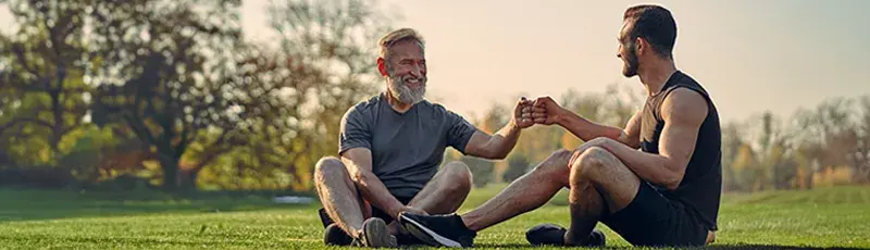 Zwei Freunde unterschiedlichen Alters sitzen mit Sportgewand im Gras und freuen sich über ihre Leistungen