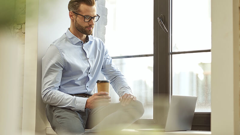 Mann mit Hemd und Brille sitzt am Fensterbrett und blickt in Laptop