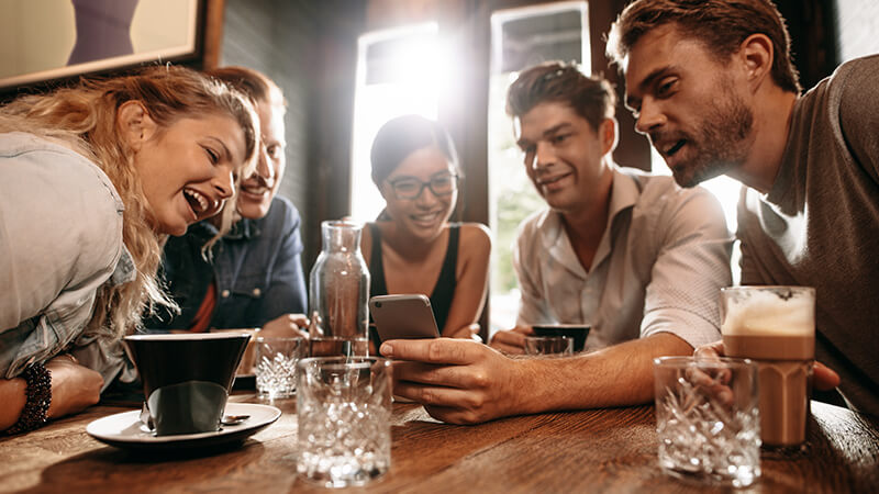 Zwei Frauen und zwei Männer sitzen mit Getränken am Tisch und blicken lächelnd in ein Smartphone