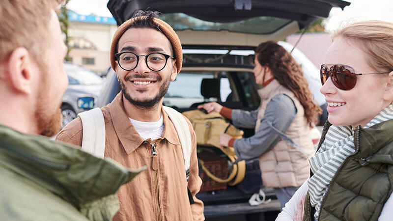 Drei Freunde unterhalten sich über ihren Roadtrip. Im Hintergrund packt eine Frau ihre Tasche in den Kofferraum.