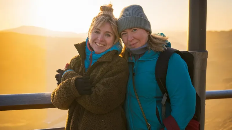 Zwei Freundinnen posieren in Winterkleidung auf einer Aussichtsplattform. Hinter den beiden sind eine weite Landschaft und der Sonnenuntergang.