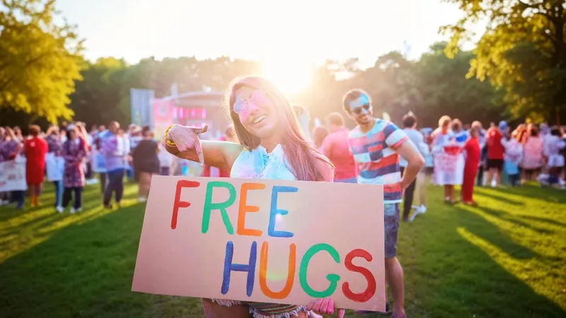 Eine Frau mit Sonnenbrille steht auf einer Wiese mit anderen Leuten im Hintergrund und hält ein buntes Schild mit der Aufschrift “Free Hugs” in der Hand