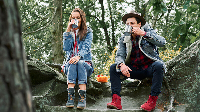 Zwei Wanderer machen eine Pause im Wald, trinken aus Eisen-Tassen und essen Obst.