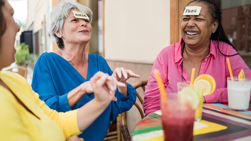 Drei Frauen sitzen lachend in einem Café und spielen Wer bin ich