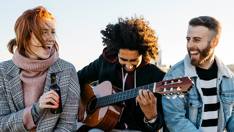 Drei Freunde sitzen lachend am Strand mit Gitarre