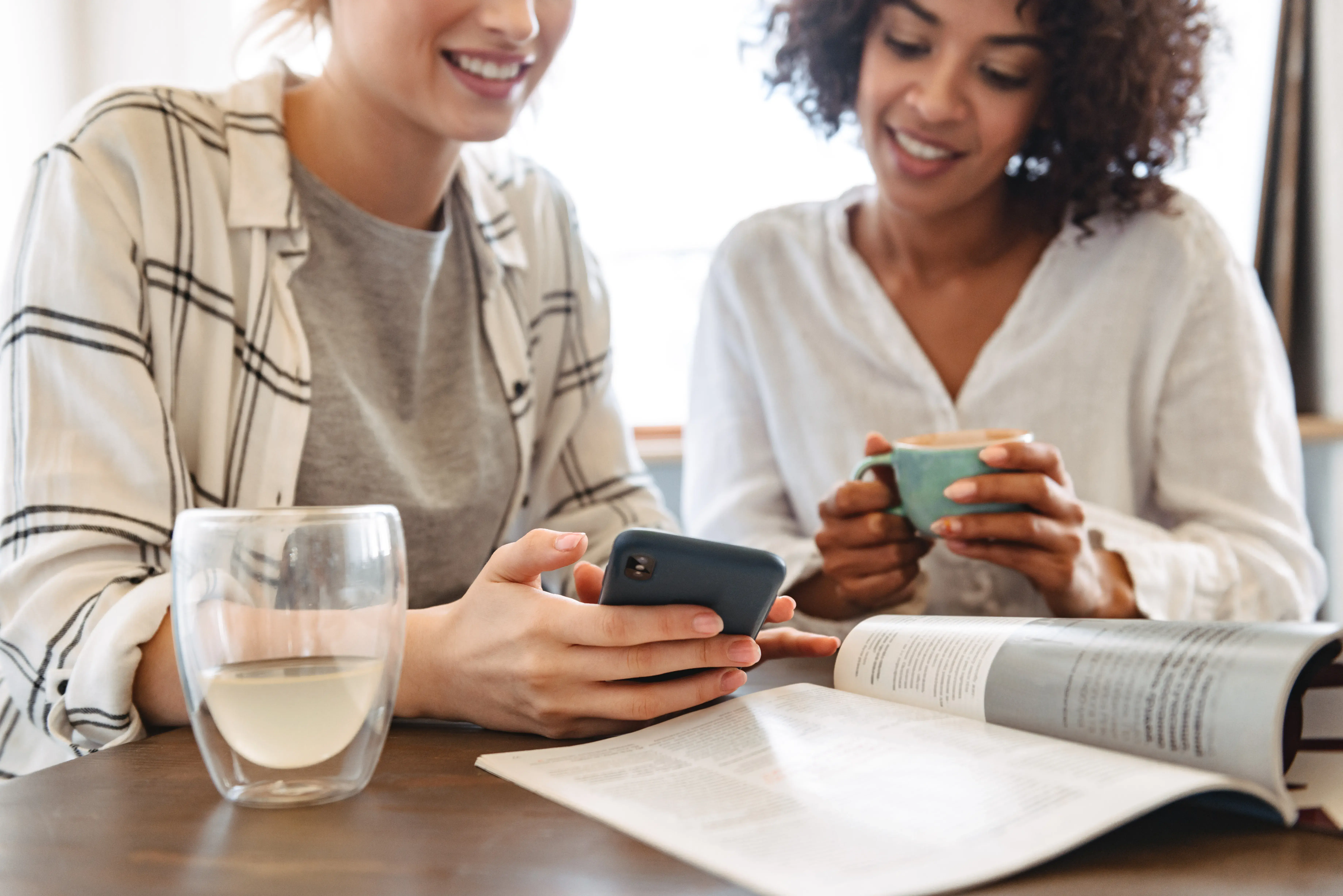 zwei Frauen sitzen am Tisch, trinken Kaffee und Tee und sehen sich eine App am Smartphone an