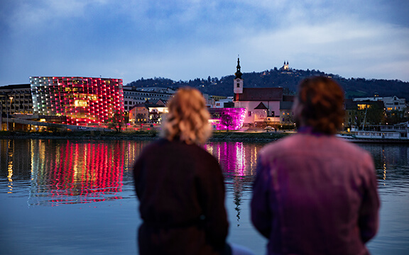 Zwei Freunde sitzen am Donauufer und blicken auf das Ars Electronica in Linz